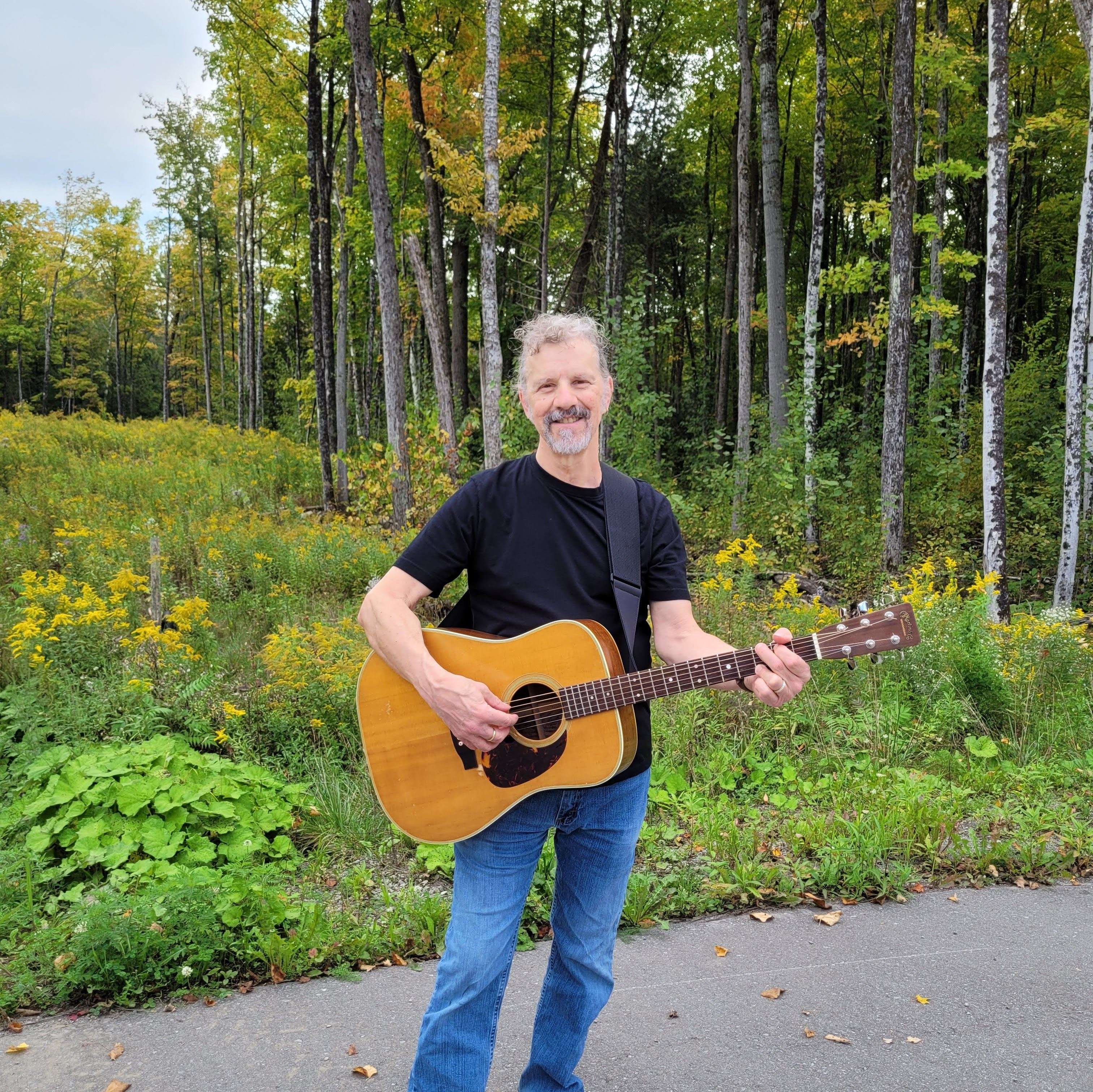 a man holding an acoustic guitar in a wooded area