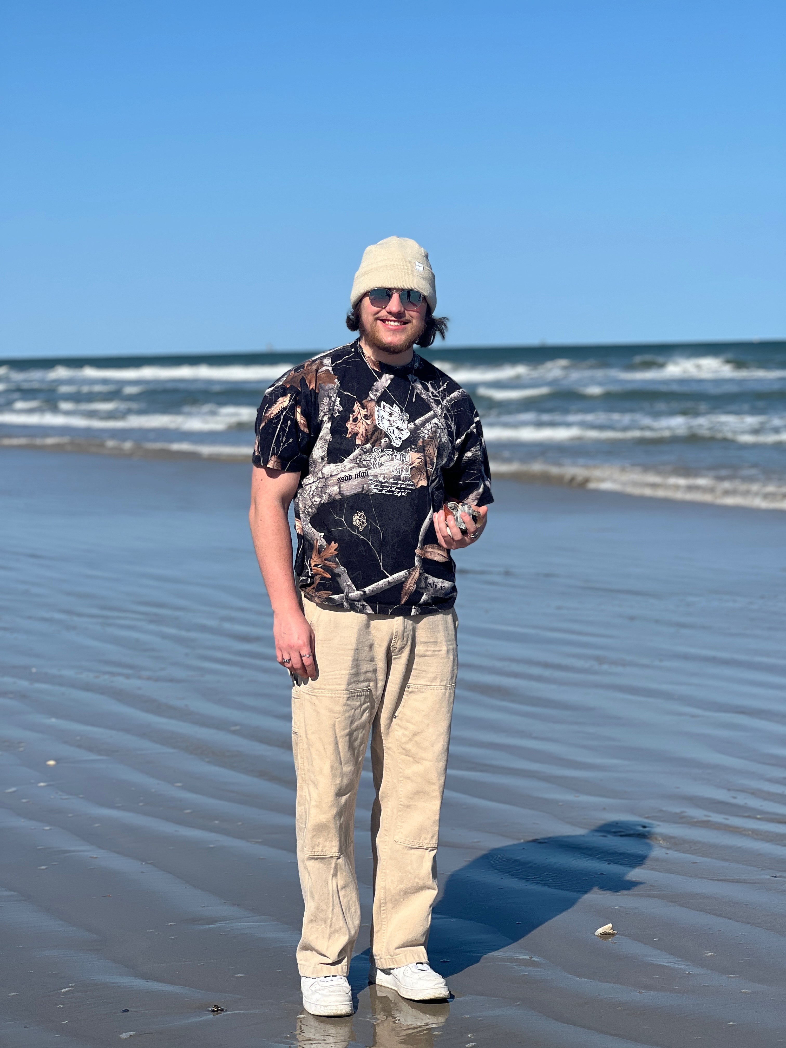 a man standing on the beach wearing a hat and tan pants