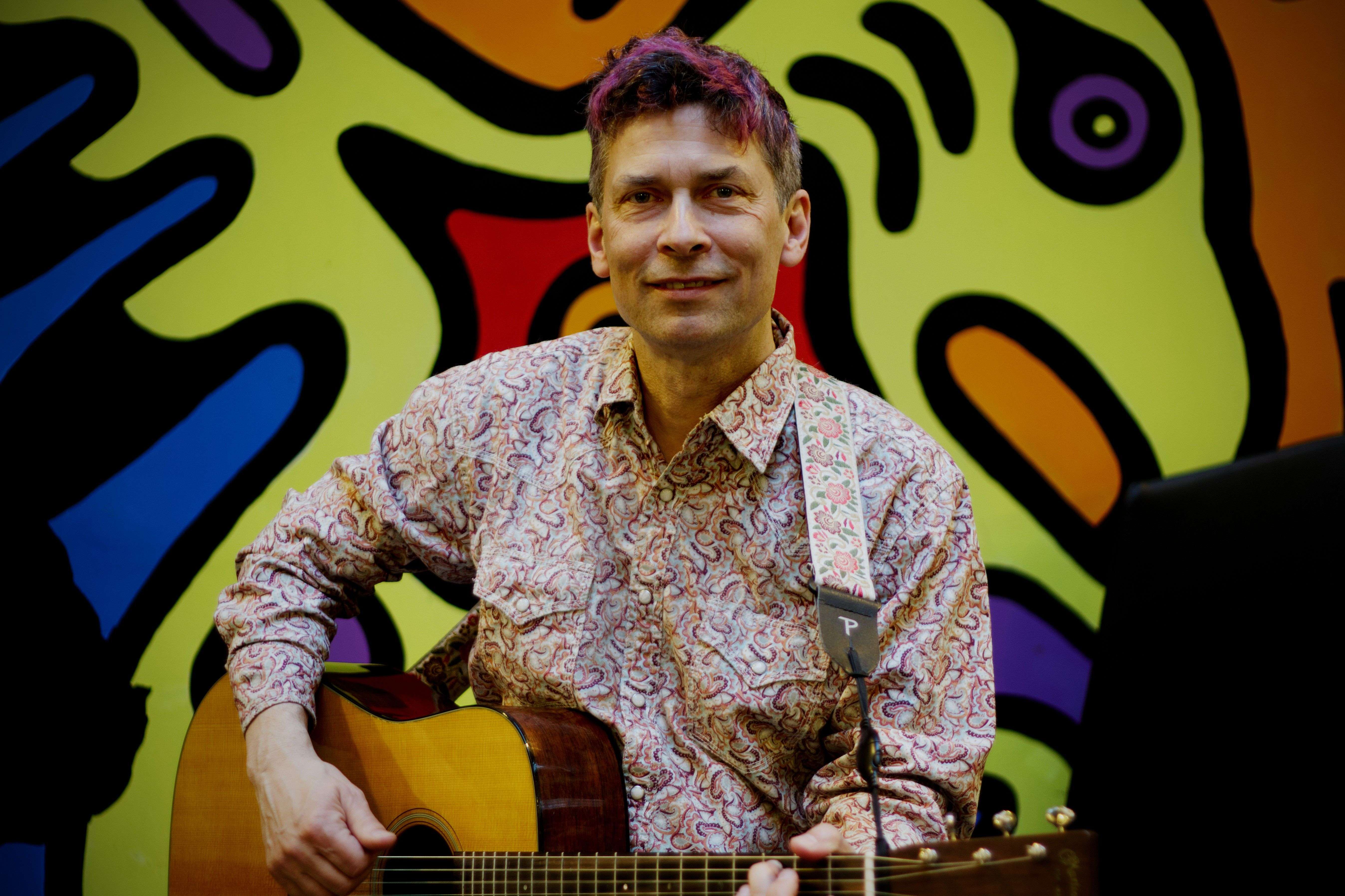 a man holding an acoustic guitar in front of a colorful wall