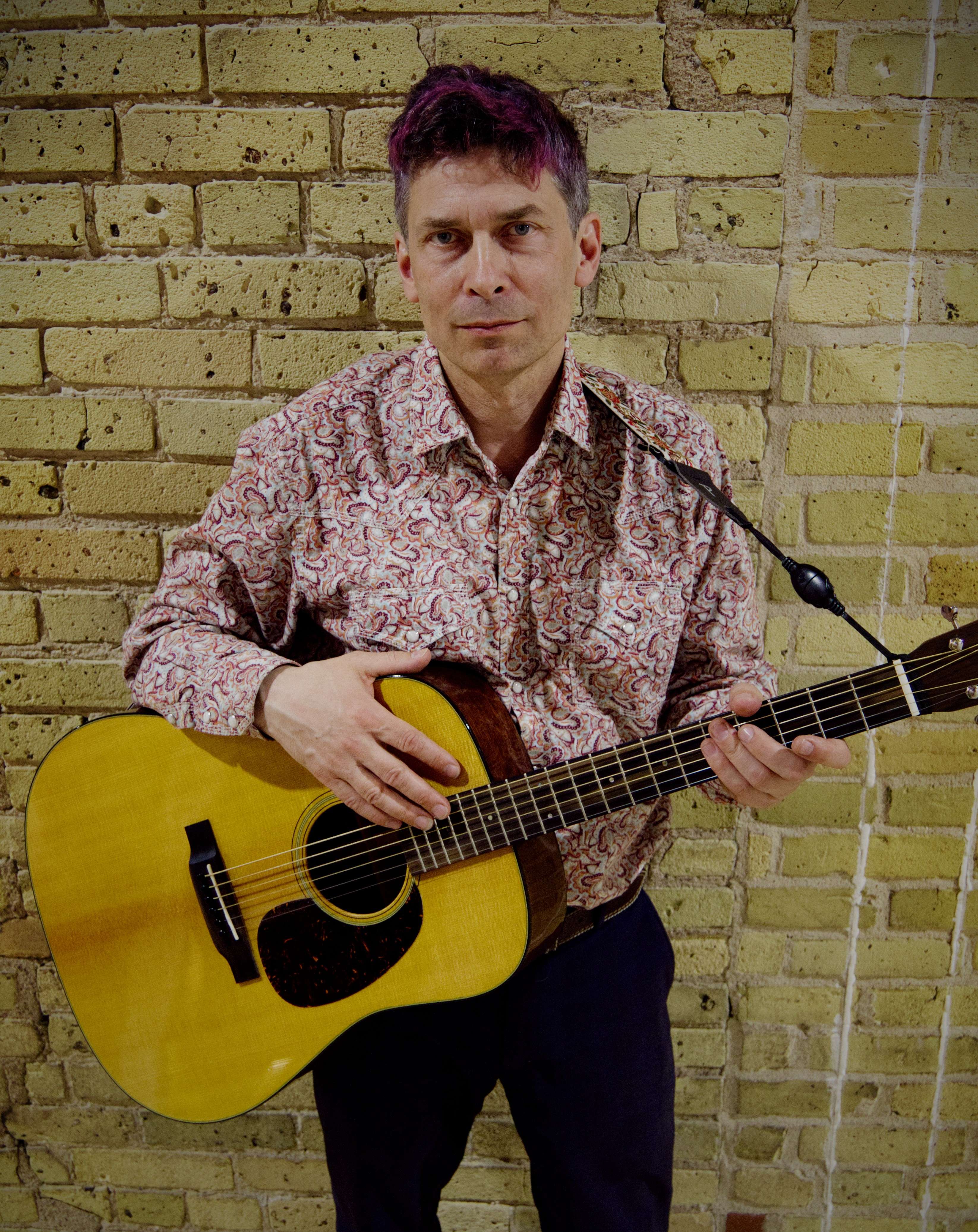 a man holding an acoustic guitar in front of a brick wall