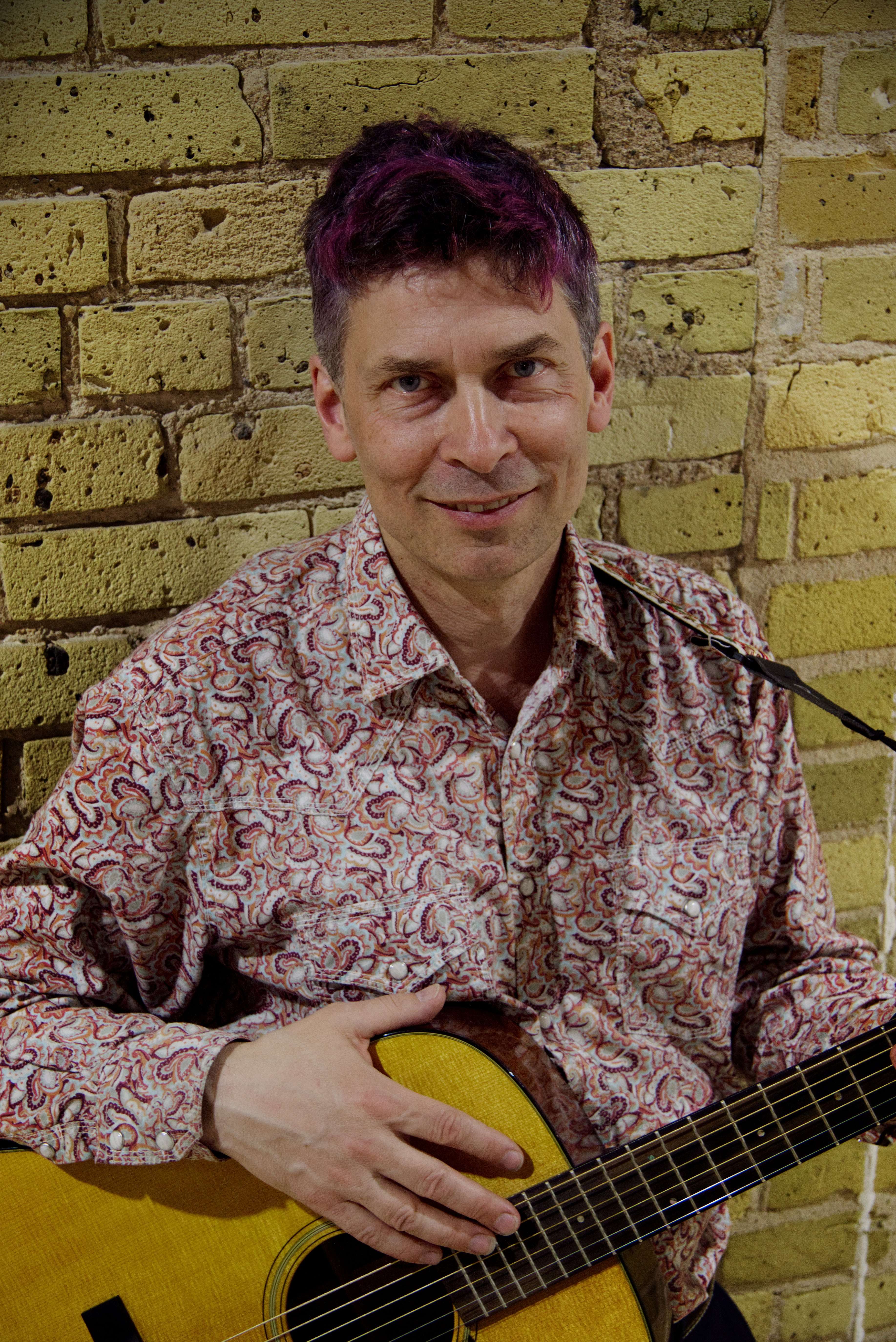 a man holding an acoustic guitar in front of a brick wall