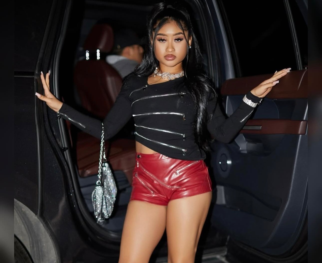 Lyric Michell (aka Young Lyric) tip jar: a woman in a black top and leather shorts standing next to a car