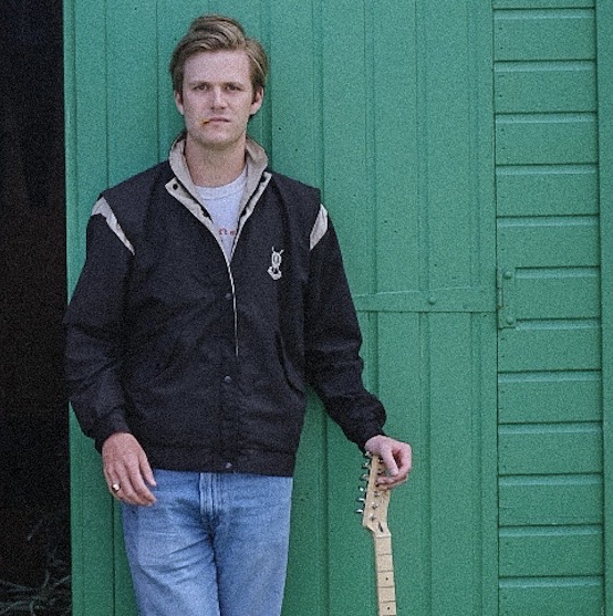 a man leaning against a green shed with a guitar