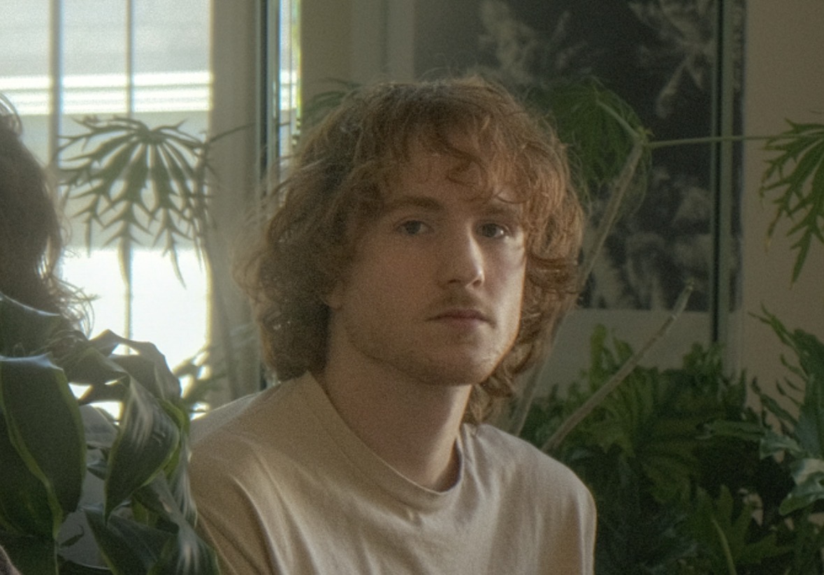 a young man with curly hair sitting in front of a plant