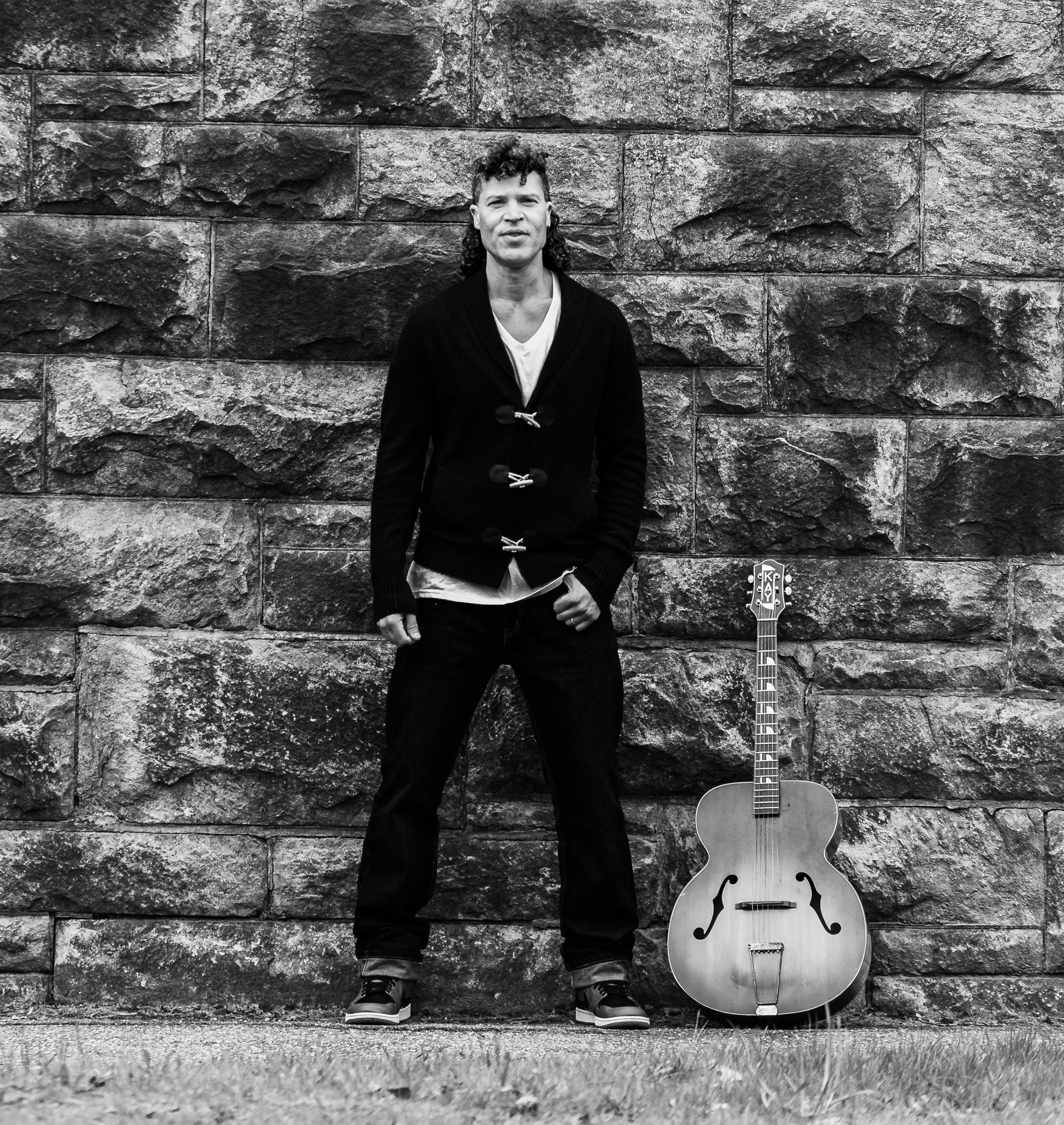 a man leaning against a stone wall with an acoustic guitar