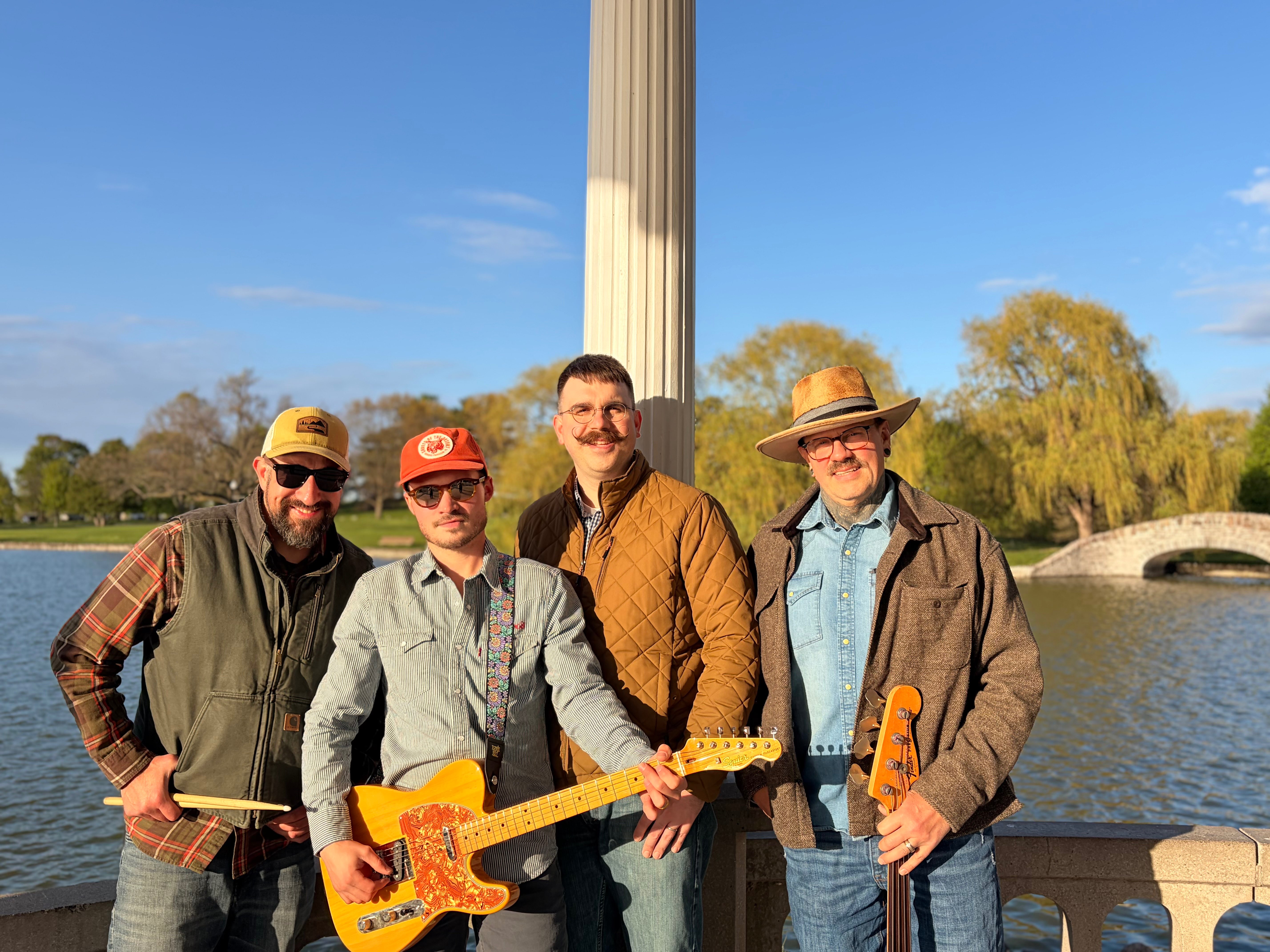 four men standing next to a pond with guitars