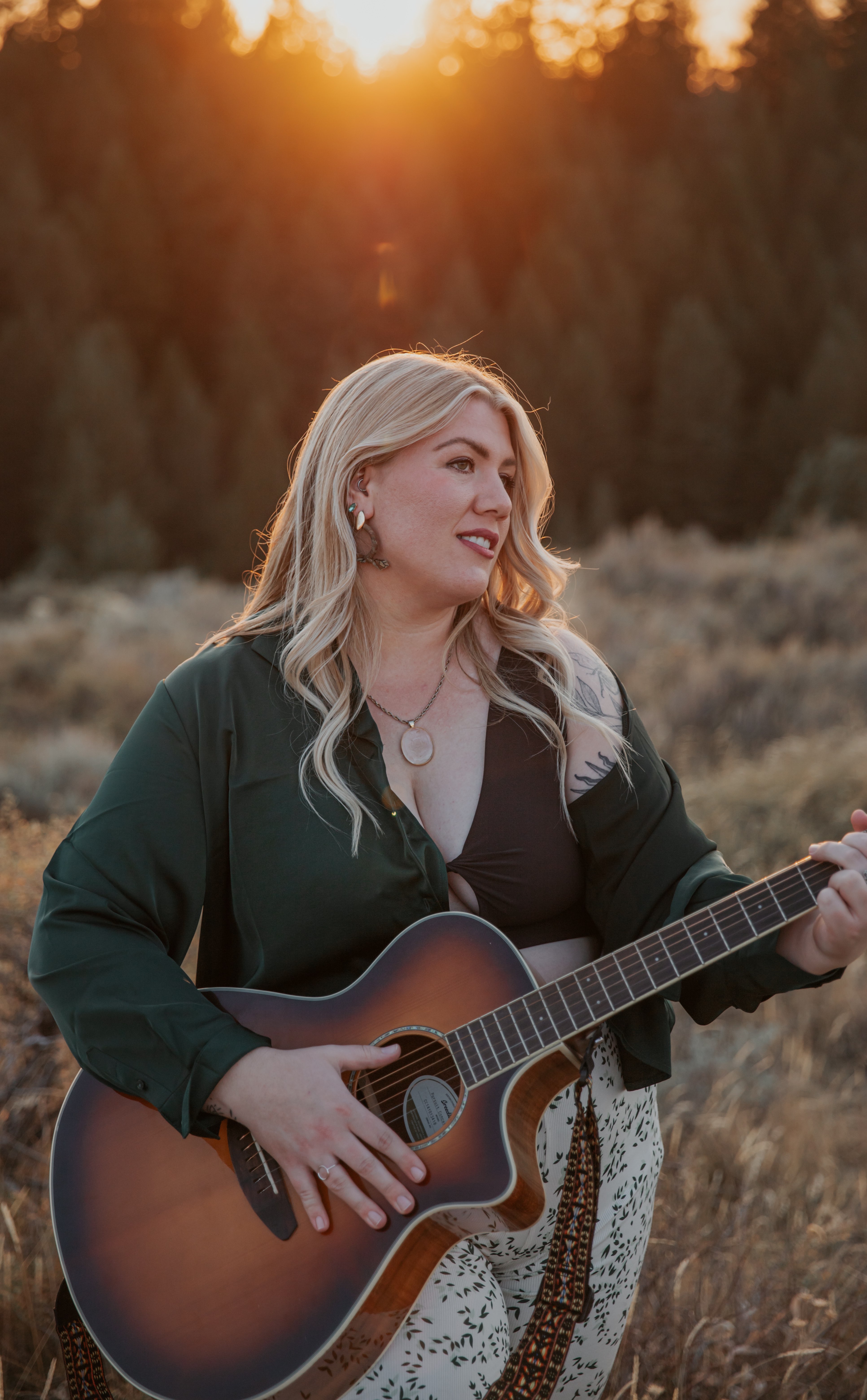 a blonde woman holding an acoustic guitar in a field at sunset