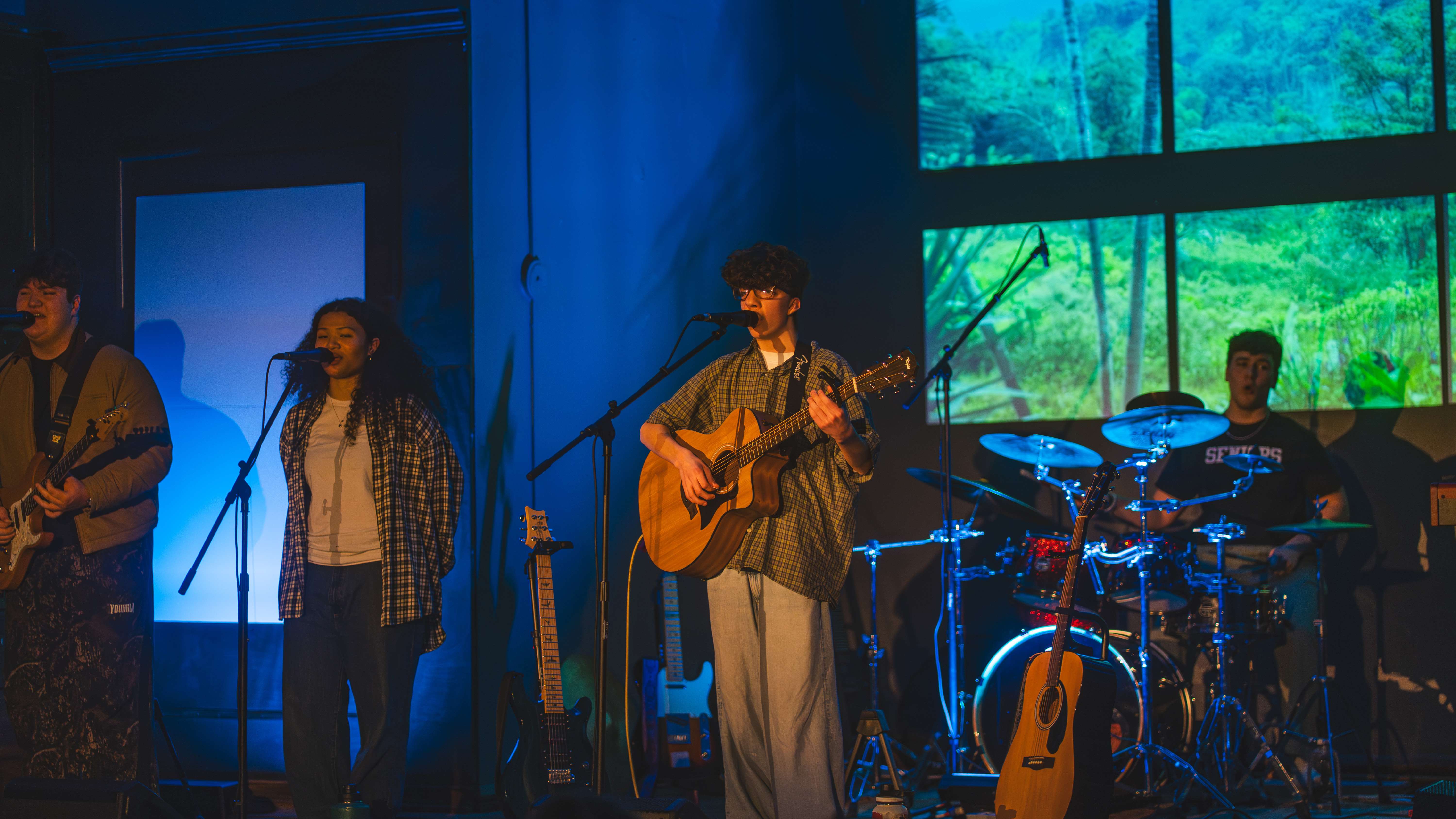 a group of people singing and playing instruments in front of a large screen