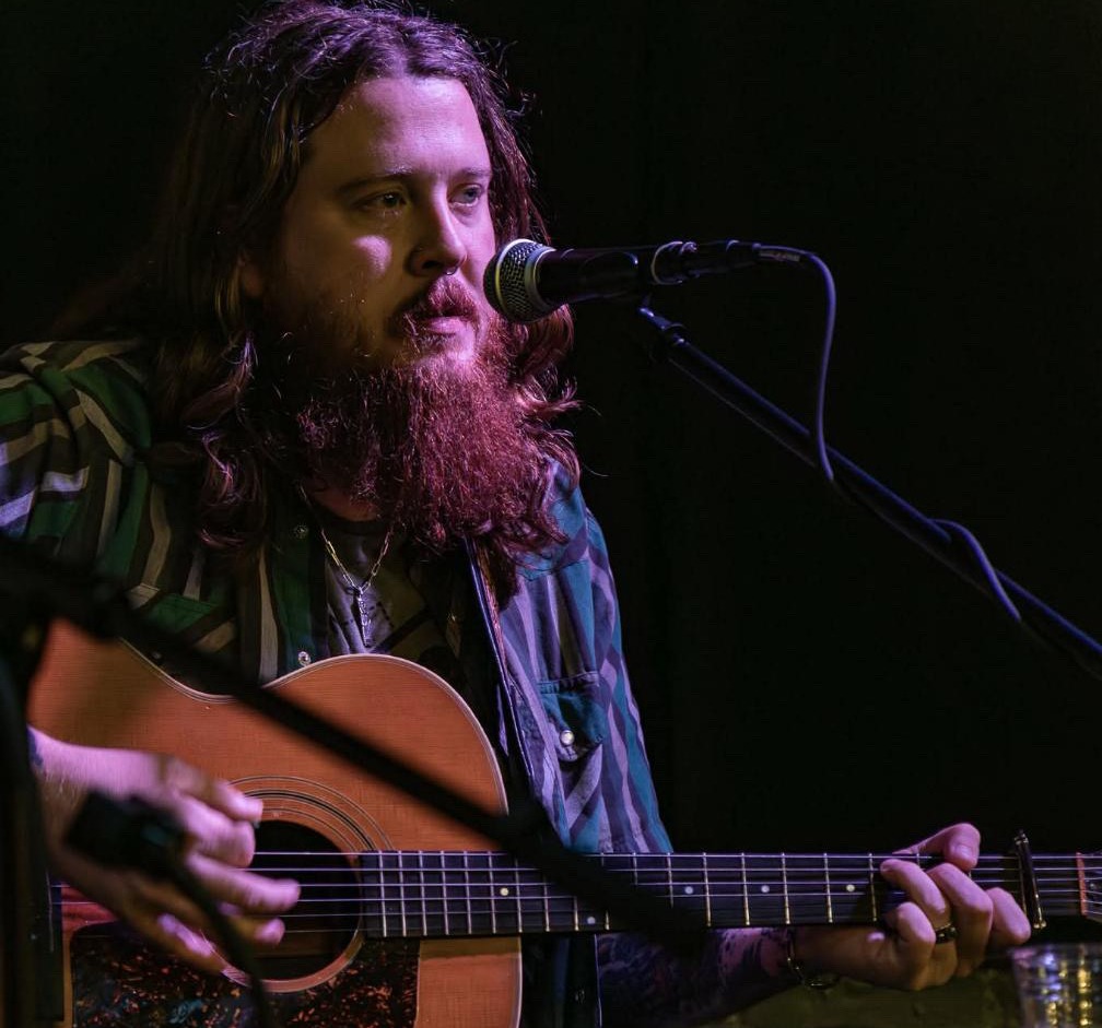a bearded man playing an acoustic guitar in front of a microphone