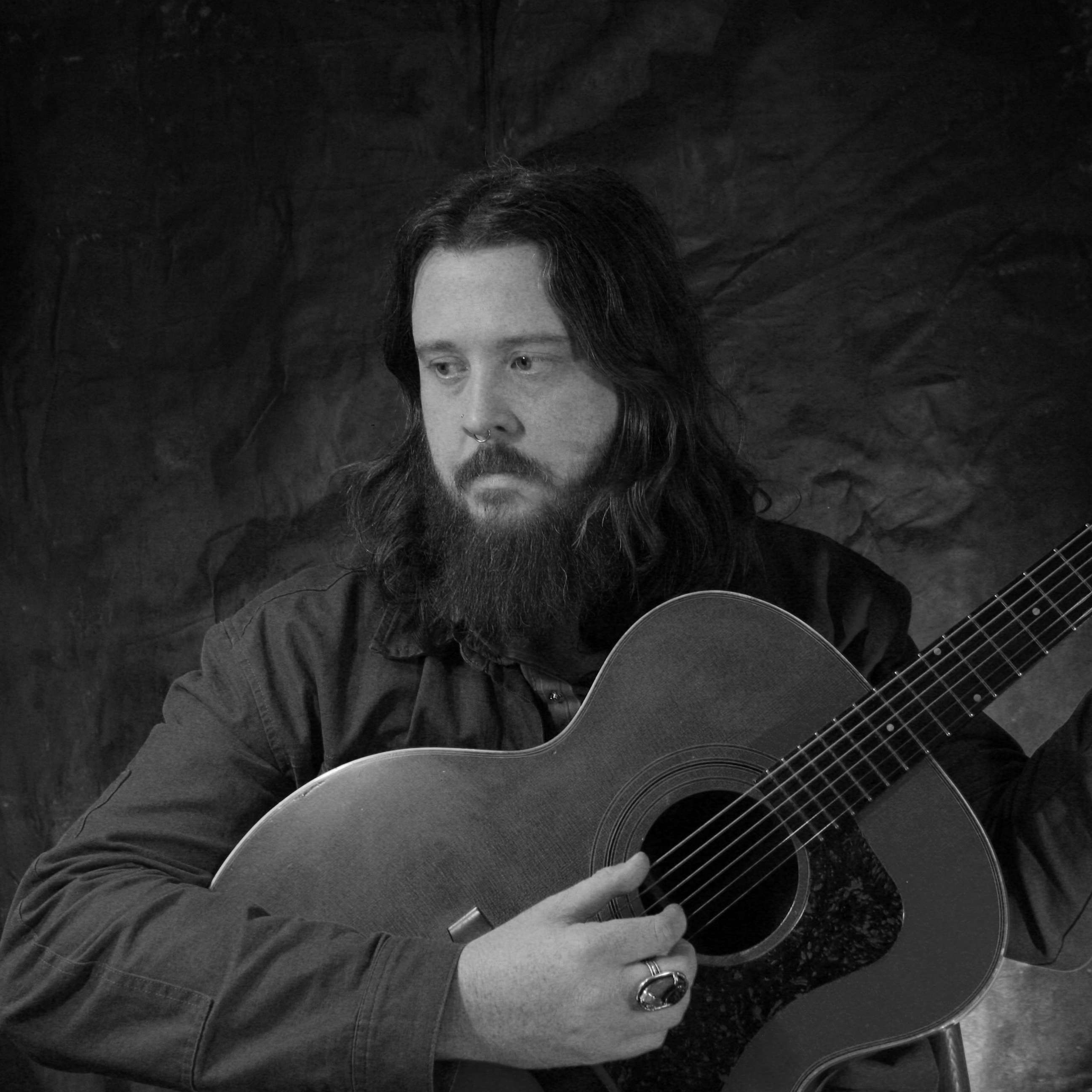 Cody Landress-Gibson - At Least Tomorrow: a bearded man holding an acoustic guitar