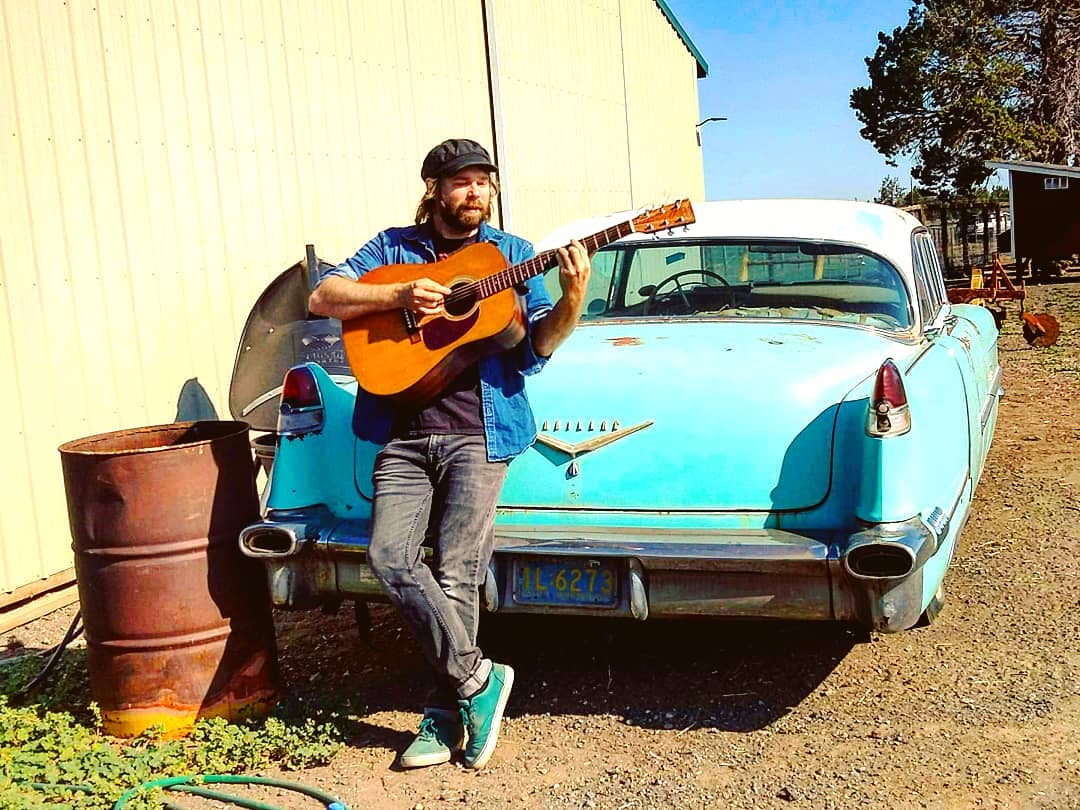 a man standing next to a blue car with an acoustic guitar
