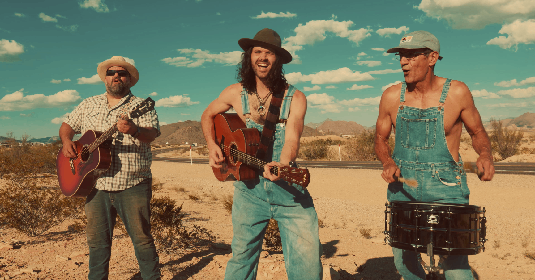 three men in overalls playing guitars in the desert