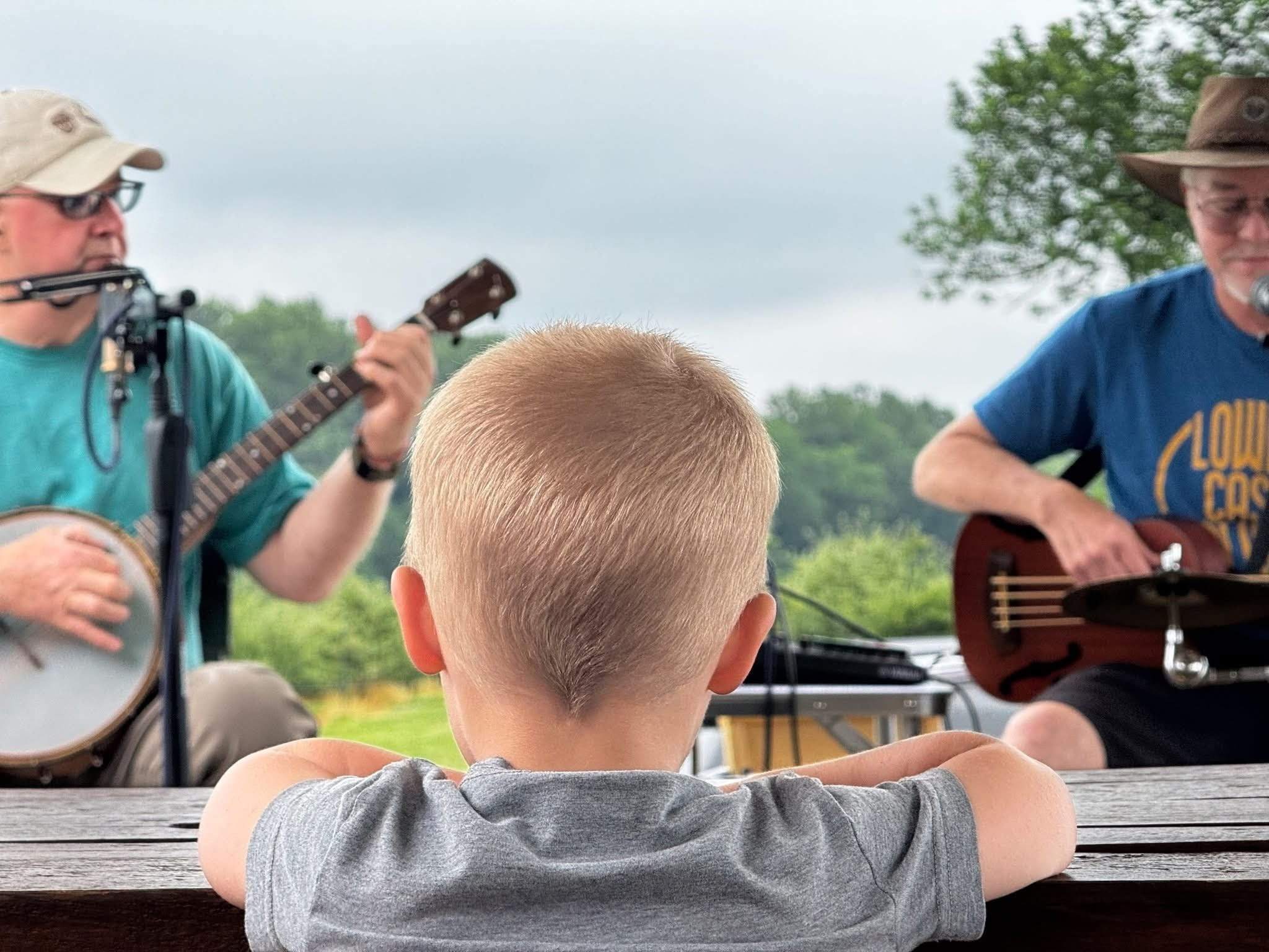 a young boy sits at a picnic table while two men play banjos